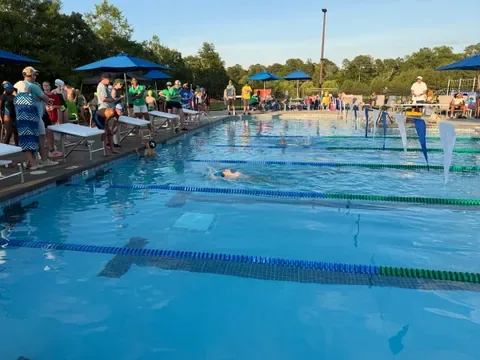 Swimmers race in the competition pool while families and swimmers gather along the deck under blue umbrellas in the evening sun at the Brightleaf Bull Sharks meet.