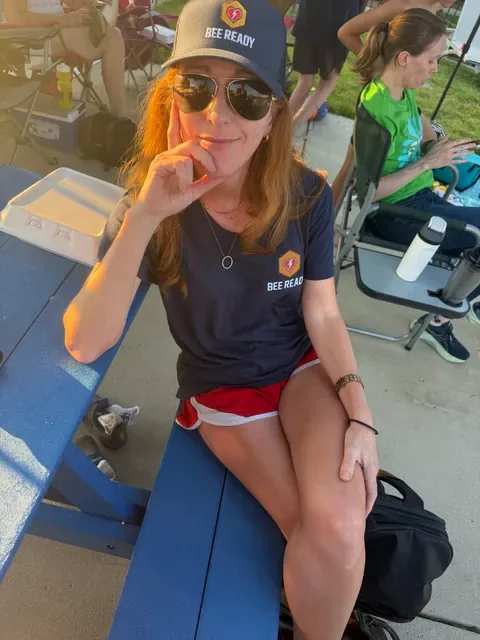 A Bee Ready volunteer in a Bee Ready cap and shirt smiles while seated at a poolside picnic table in the golden evening light.