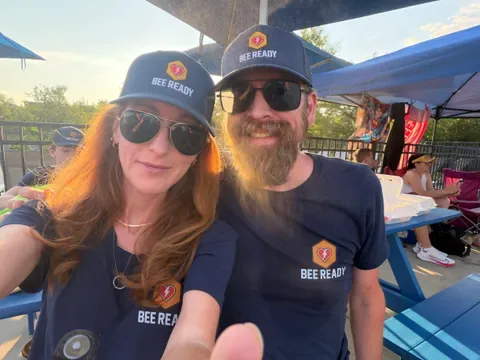 Two Bee Ready volunteers in navy Bee Ready caps and shirts smile for a poolside selfie at the swim meet.