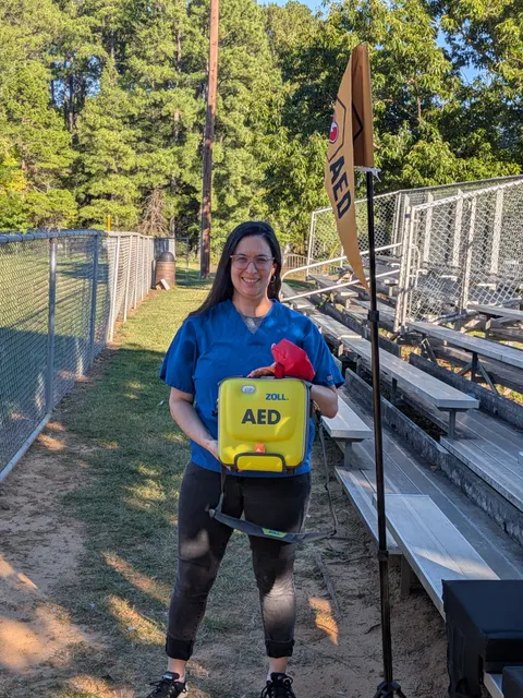 La voluntaria de BeeReady Aviva Emmons, con uniforme médico azul, sonríe mientras sostiene un DEA amarillo junto a las gradas y una bandera de DEA de BeeReady en el partido de South Durham Little League.