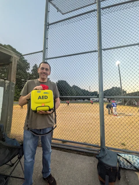 BeeReady volunteer Laney Emmons holds a yellow AED beside the backstop at a youth baseball game.